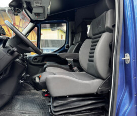 Inside a blue truck cab: the gray driver's seat with ribbed back, large steering wheel, dashboard and overhead storage pockets, and a textured rubber floor mat visible; passenger seat in the background.