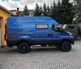 Blue high-roof camper van parked on a paved driveway beside a yellow building, with another camper in the background.