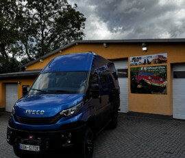 Blue Iveco van parked in a paved lot outside a yellow-orange workshop with wall posters behind it.
