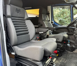 Interior of a blue truck cab with two gray ergonomic seats, armrests, and a rugged rubber floor mat.