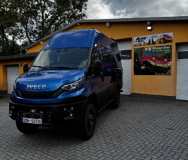 Blue Iveco van parked on a brick driveway in front of an orange building with an off-road poster on the wall.